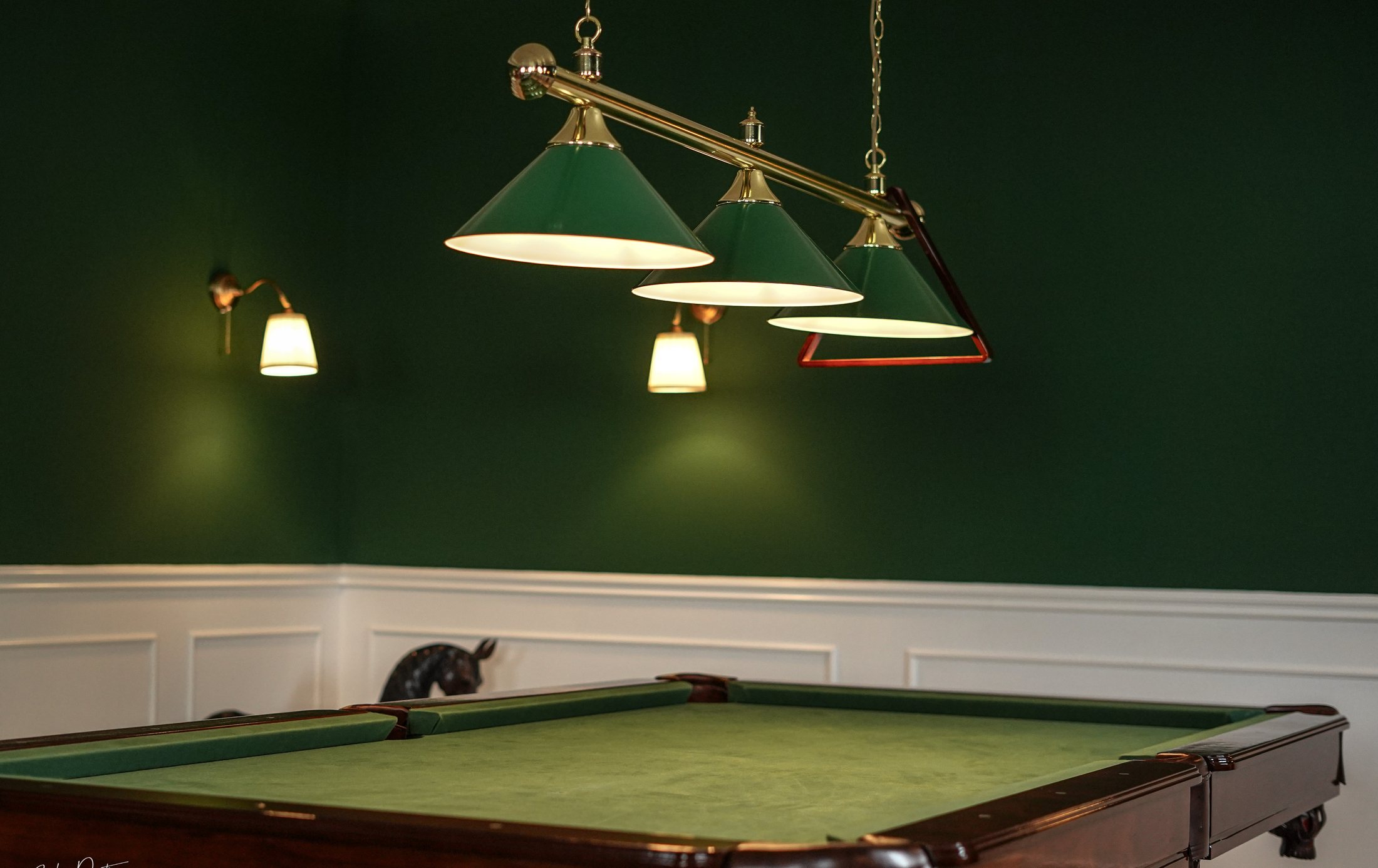 Green pendant lamps above a billiard table, bronze horse behind.