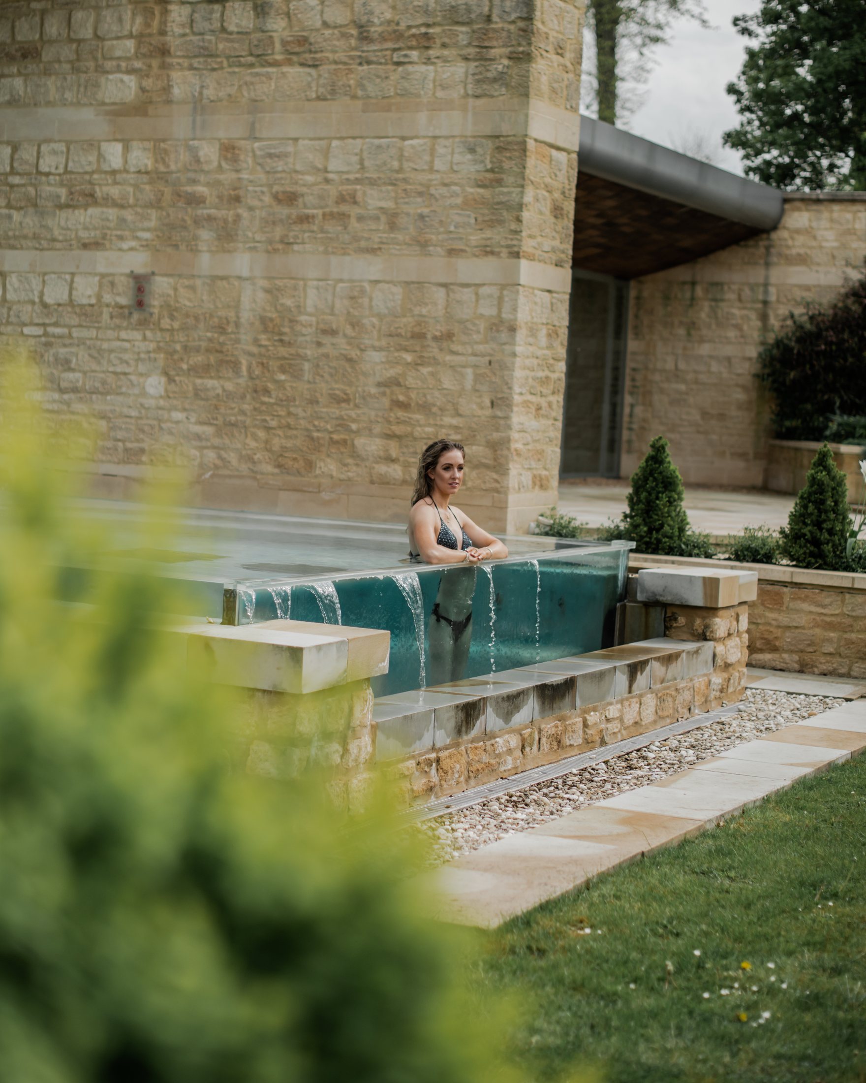 The guest in the outdoor plunge pool, water spilling over the glass edge against Cotswold stone.